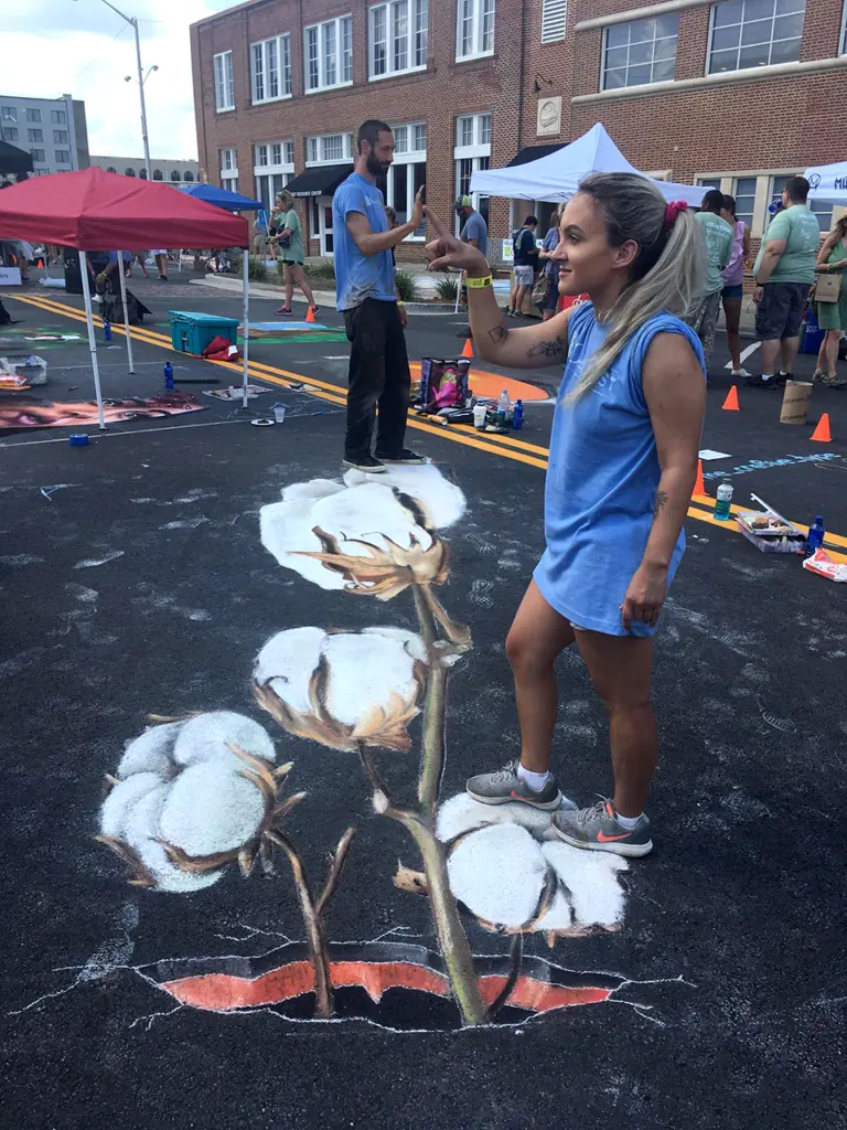 A woman stands on a 3D chalk drawing of a cotton plant that appears to grow up out of a hole in the street.