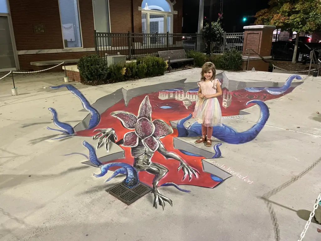 3D chalk drawing of a petal-faced monster climbing out of a red crack in the pavement with blue tentacles reaching out, as a young girl in a pink dress stands at its edge.