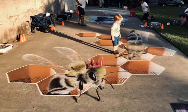 3D chalk drawing of a giant crowned honeybee climbing out of an orange honeycomb, with a young child standing among the cells.