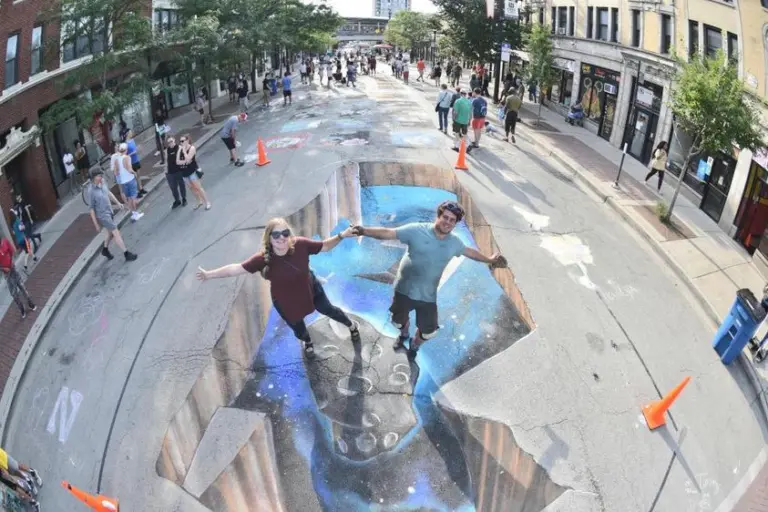 Two people pose with arms outstretched inside a 3D chalk illusion of a deep chasm that opens into outer space in the middle of a city street.