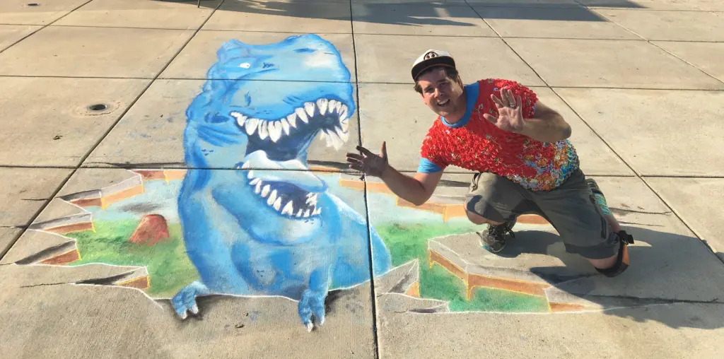 Zach crouches and gestures beside his 3D chalk drawing of a blue T. rex that appears to burst out of the sidewalk with its mouth wide open.