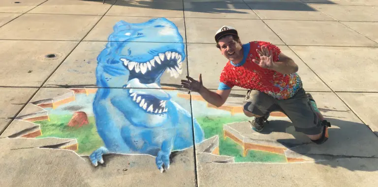 Zach crouches and gestures beside his 3D chalk drawing of a blue T. rex that appears to burst out of the sidewalk with its mouth wide open.
