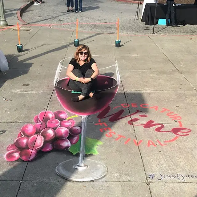 A woman sits inside a giant 3D chalk wine glass surrounded by grapes and Decatur Wine Festival lettering.