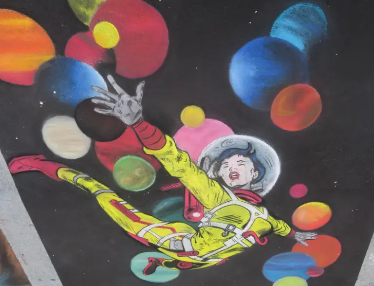 Chalk drawing of an astronaut in a yellow suit floating among colorful planets on a black background.