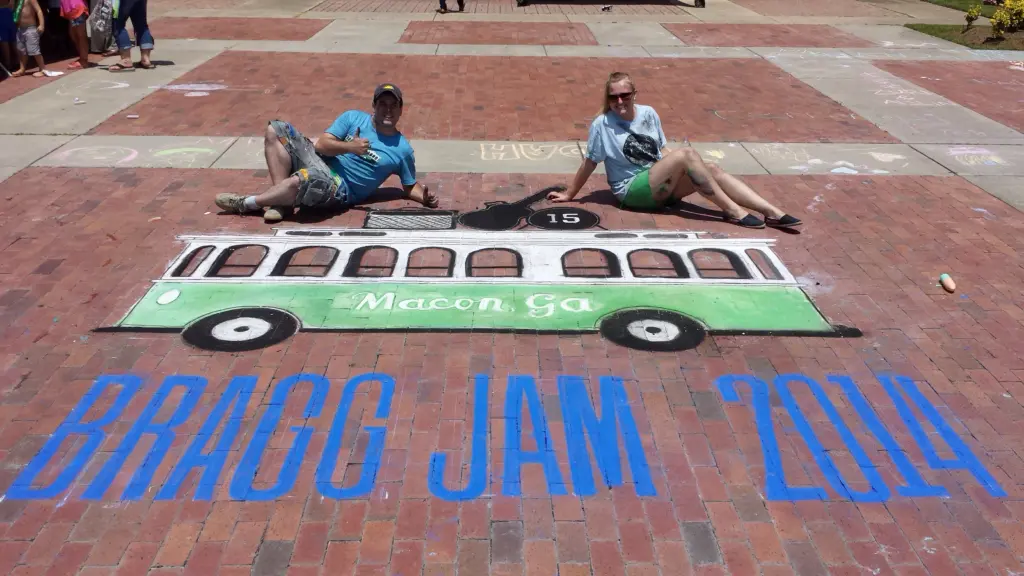 Zach and Jessi Queen lie on either side of their chalk drawing of a green Macon trolley above large blue letters reading Bragg Jam 2014.