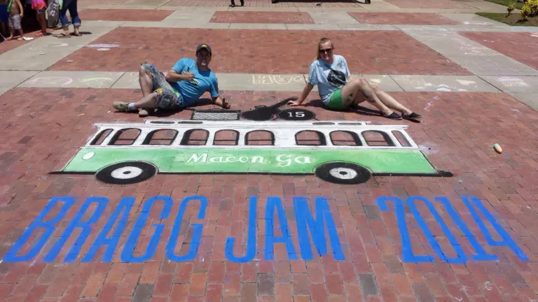Zach and Jessi Queen lie on either side of their chalk drawing of a green Macon trolley above large blue letters reading Bragg Jam 2014.