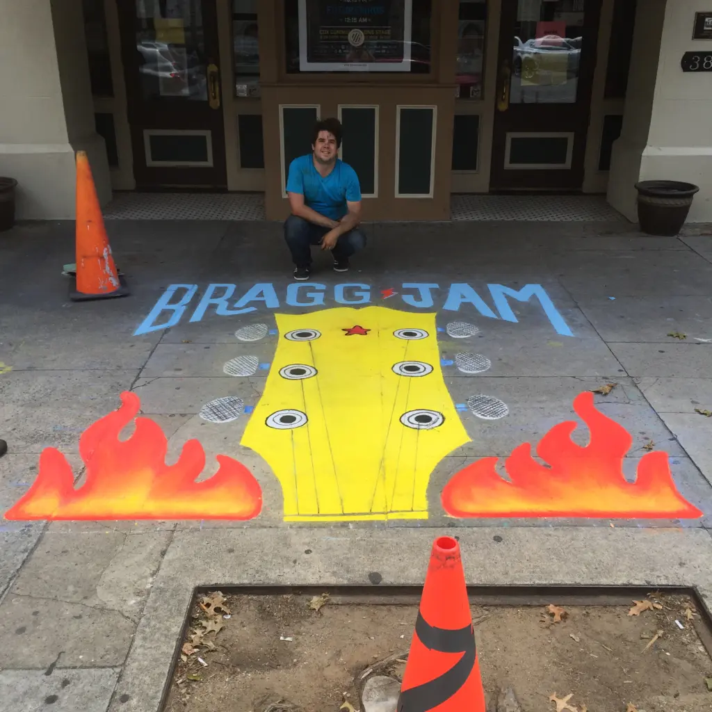 Bragg Jam street painting of a yellow guitar headstock between two orange flames, with Zach sitting behind it in front of a venue door.