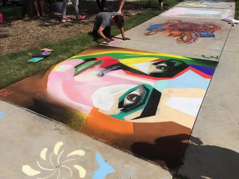 Colorful chalk face in a cubist style, built from bold blocks of pink, green, yellow and black on a sidewalk, with other artists working in the background.