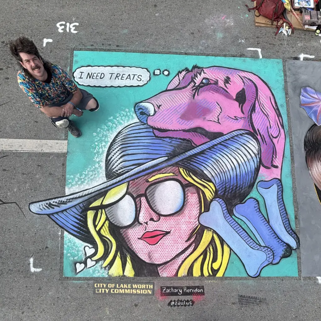 Zach Herndon kneels beside his pop-art chalk drawing of a woman in a wide sun hat and sunglasses, with a pink dog above her thinking "I need treats" and blue dog bones around her.