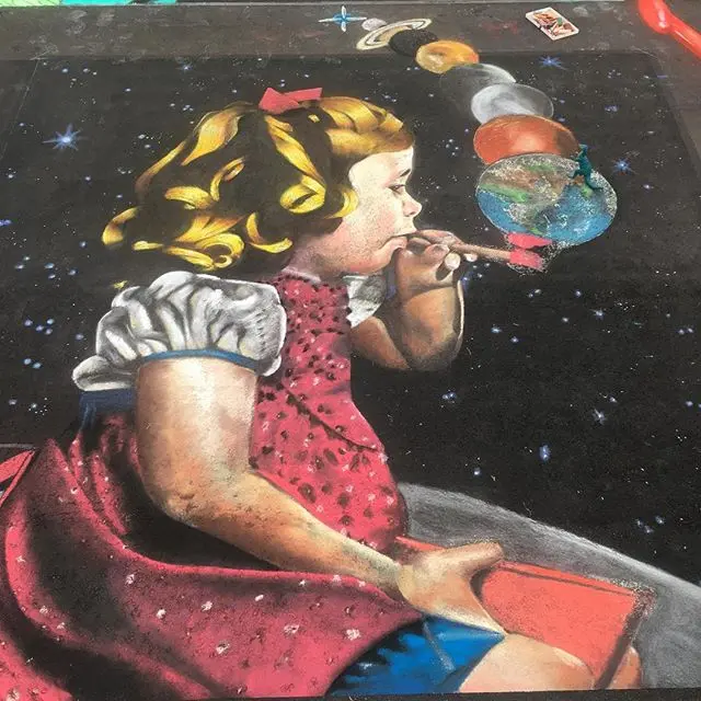 Chalk painting of a little girl in a red dress blowing bubbles that turn into planets against a black sky.