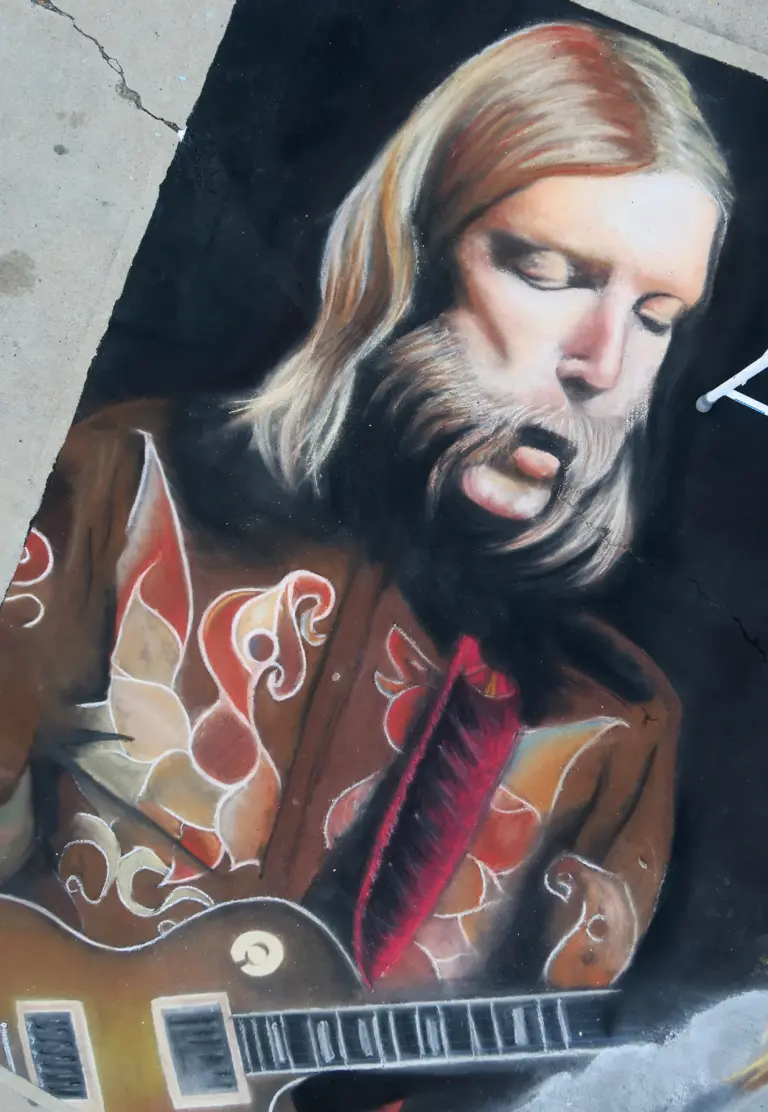 Chalk portrait of a long-haired, bearded musician playing an electric guitar, wearing an embroidered jacket in red and orange.