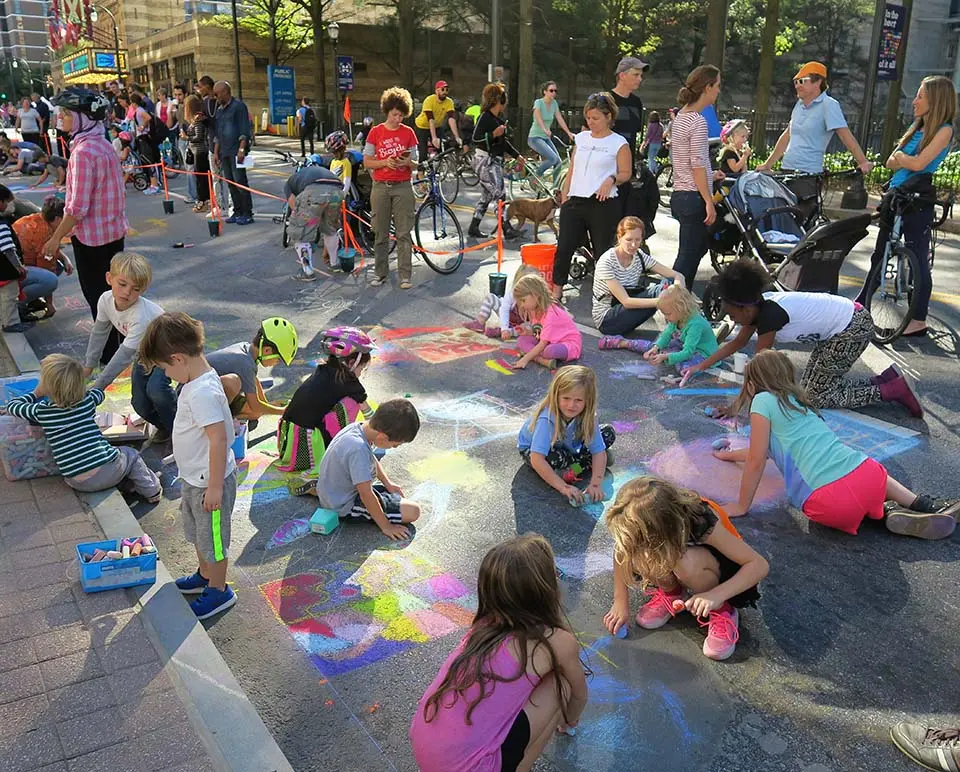 Children and adults kneel side by side on Peachtree Street, coloring chalk squares at a Pop-Up Chalk Fest.