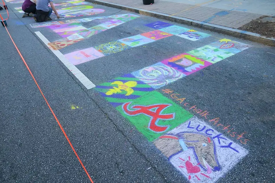 Rows of chalk squares on a closed street, each colored by a different visitor as part of one larger community design.
