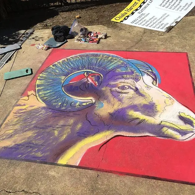 Chalk drawing of a ram's head with curling horns in purple and gold on a red background, with chalk supplies at the edge.