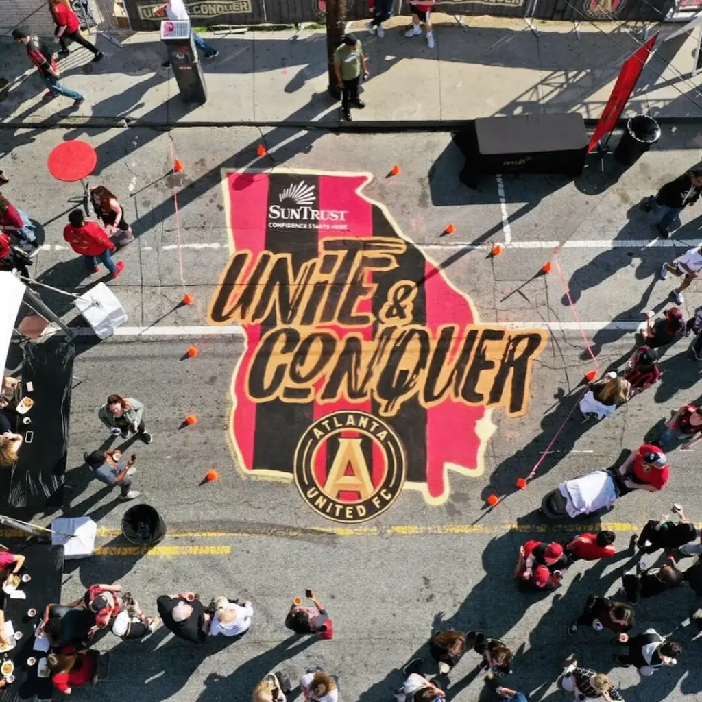 Overhead view of a large street mural shaped like the state of Georgia in red and black stripes, reading Unite and Conquer above the Atlanta United crest, with fans gathered around it.