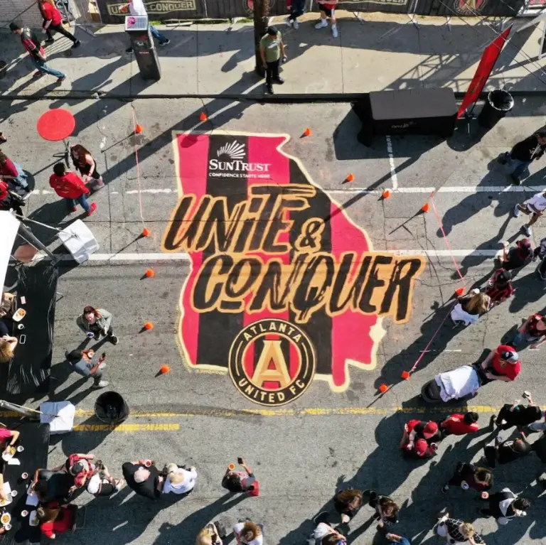 Overhead view of a large street mural shaped like the state of Georgia in red and black stripes, reading Unite and Conquer above the Atlanta United crest, with fans gathered around it.