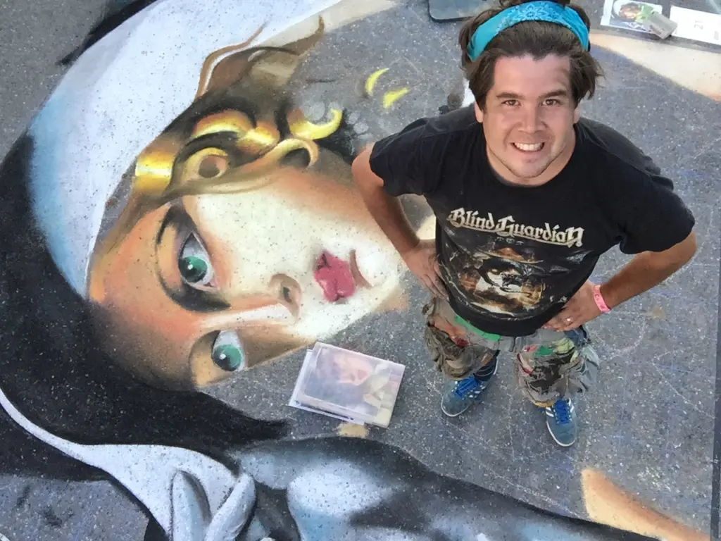 Zach Herndon grins up at the camera, standing beside his large chalk portrait of a woman with green eyes and a gold headpiece.